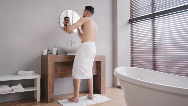 Full length view funny cheerful man with white bath towels wrapped around hips standing in bathroom looking in mirror naked guy dancing after shower having fun moving to music happy dance moving body — Vídeos de Stock