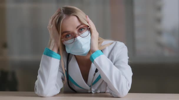 Portrait tired exhausted girl nurse female doctor pediatrician cardiologist ophthalmologist dentist gynecologist feels stress pressure wears glasses medical gown protective face mask sitting at table — стоковое видео