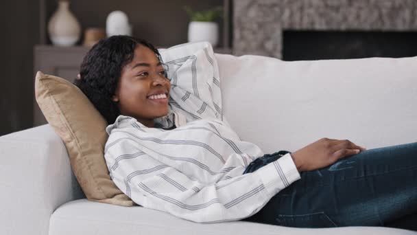 Happy relaxed african american woman rest lounge lying on couch enjoy peaceful mood weekend casual rest in living room on sofa lazy calm girl dreaming smiling on comfortable furniture cozy day at home — ストック動画