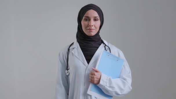 Young arabian muslim woman islamic doctor surgeon in hijab wear medical white coat stethoscope looking at camera female professional physician stand posing in clinic office holding folder documents — стоковое видео