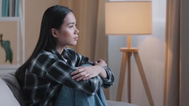 Вид сбоку грустная азиатская женщина сидит на диване дома, чувствуя себя неуверенным в трудном решении несчастливое плохое чувство психические проблемы сидит на диване. Депрессивная корейская леди сожалеет об ошибке психологической проблемы — стоковое видео