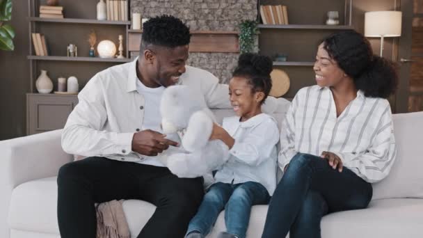 African happy family parents with child daughter sitting on sofa at home having fun sincerely rejoice child with teddy bear toy playing laughing enjoying play game with mother and father at home couch — ストック動画