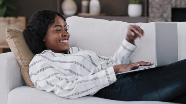 Χαμογελώντας αφρικανή φοιτήτρια ελεύθερη επαγγελματίας χρήστης ξαπλωμένη στον καναπέ χρησιμοποιώντας το μήνυμα δακτυλογράφησης laptop σε απευθείας σύνδεση συνομιλία με τους φίλους ευτυχής συνομιλία στην περιήγηση στον υπολογιστή στο διαδίκτυο social media freelance work — Αρχείο Βίντεο