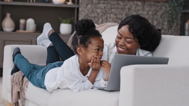 Happy parent african mother and cute child daughter looking at laptop screen talking having fun video call choosing in internet store using computer app game at home relaxing laughing lying on sofa — Video Stock