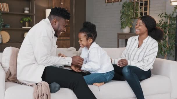 Happy african family mum dad with cute little kid daughter tickling having fun relax on sofa together laughing young carefree parents and child girl laugh play cuddling at home on couch casual game — Video