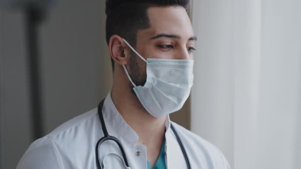 Male pensive thoughtful arab man therapist surgeon wear white uniform medical face mask standing at workplace looking out window preventing spread infection pandemic outbreak close-up doctor portrait — Vídeos de Stock