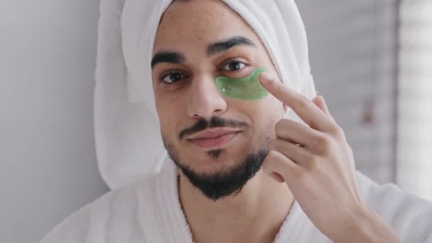 Забавный портрет мужского лица улыбающийся индийский арабиец, одетый в полотенце для ванны на голове клей липкий гидрогель для глаз косметические процедуры по уходу за кожей парень смотрит в камеру с коллагеновыми пятнами под глазами — стоковое видео