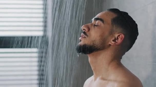 Portrait Arab Spaniard Indian young handsome manly wet man bearded brunette guy washes in shower enjoys falling water flowing over male face washing hair refreshed after workout in gym morning hygiene — Video