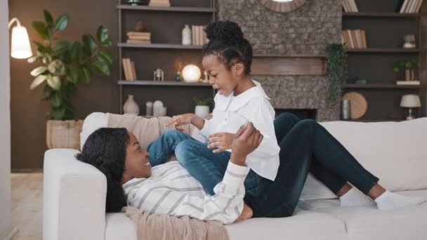 Szczęśliwy relaks afrykańska matka leżąca na kanapie i małe dziecko aktywne zabawy córka dziewczyna na kanapie śmiejąc się o zabawy rozmawiając rozmowa uśmiechnięta mama korzystając bonding spędzać czas z dzieckiem — Wideo stockowe
