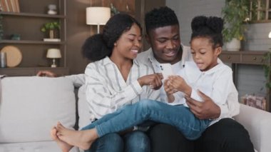 Şirin liseli kız çocuğu olan mutlu Afrikalı aile ebeveynleri kanepede oturup komik akıllı telefon uygulamaları okuyarak, internette şakalar okuyarak, telefonda eğlenerek, gülümseyerek evde cep telefonu ekranlarına bakarak eğleniyorlar.