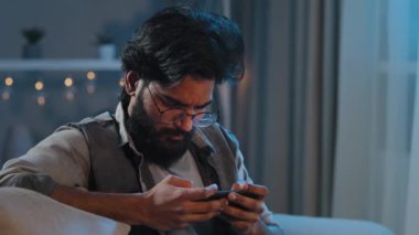 Yakın plan odaklı motivasyonlu Arap sakallı adam gözlüklü, mobil oyun oynayan online spor yarışması internet konsolu gece evde oturup akıllı telefon bağımlılığıyla oyun oynuyor.
