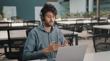 Genç Arap erkek işçi operatörü, kulaklıklı, mikrofon kullanan, dizüstü bilgisayarı kullanan, müşteriyle konuşan, profesyonel destek veren, şirket ürünü satan, web kamerasında finansal dokümanlar satan.