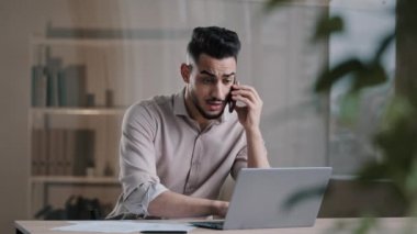 Bin yıllık Arap iş adamı web tasarımcısı cep telefonuyla konuşuyor çoklu görevlerde yer alan bilgisayarlar kullanarak online projeleri tartışıyor ofisteki kağıt dökümanlarını not ediyor.