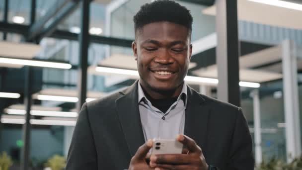 Счастливый улыбающийся африканский парень американец бизнесмен босс работник держа современное беспроводное устройство смс сообщение на смартфоне с помощью социальных медиа приложений просмотра Интернета играть мобильные игры в офисе — стоковое видео