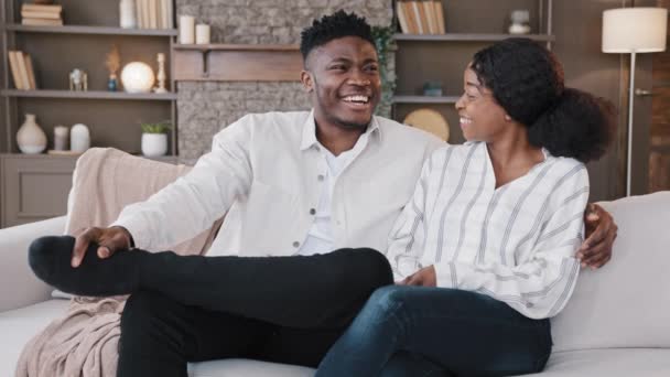 Familie koppel gehuwde echtgenoten Afrikaanse man en biracial vrouw praten lachen zitten samen op de bank in de woonkamer in nieuw huis eigen appartement liefdevolle man en vrouw hechting gesprek thuis — Stockvideo