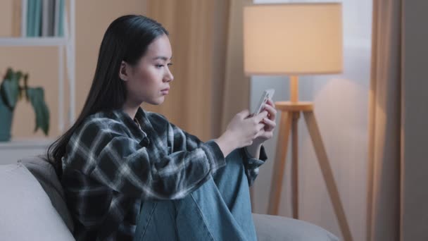 Vista lateral de triste aburrido mujer asiática chica sentada en el sofá en casa con el teléfono de la señora infeliz grave desplazamiento de navegación en el teléfono inteligente y el uso de la aplicación de teléfono móvil viendo noticias de redes sociales alimentación mecanografía — Vídeos de Stock