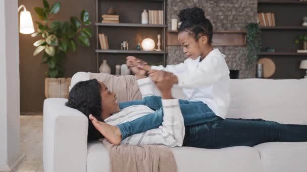 African American loving mother mommy lying on sofa relaxing at home living room with little daughter child talking having fun casual game playing holding hands baby rocking back forward sitting on mum — ストック動画