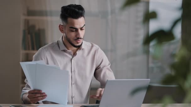 Serious arabian young business guy male professional worker employee type on computer keyboard copy paper text at electronic form focused hispanic man compare documents check online financial report — Vídeo de Stock