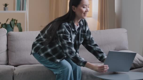 Carefree Asian girl enters living room sit down on comfortable sofa takes laptop with smile and watches video online woman uses computer sitting on couch at home casual browsing net resting typing — стоковое видео