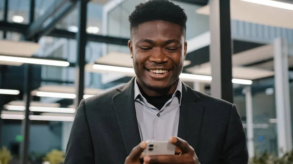 Mutlu gülümseyen Afrikalı adam Amerikalı işadamı patron modern kablosuz cihaz sahibi sosyal medya uygulamaları kullanarak akıllı telefondan mesaj yolluyor ofisteki mobil oyunlar oynuyor. — Stok fotoğraf