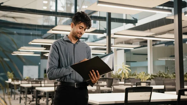 Young arabian confident business man guy stand in modern office hold folder check document male hispanic entrepreneur accountant read financial report analyze audit papers satisfied looking at camera — стоковое фото