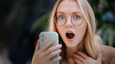 Portrait caucasian girl blonde successful lucky woman in glasses looks at screen of mobile phone wins online message with offer invitation happiness victory shock emotion good news with smartphone
