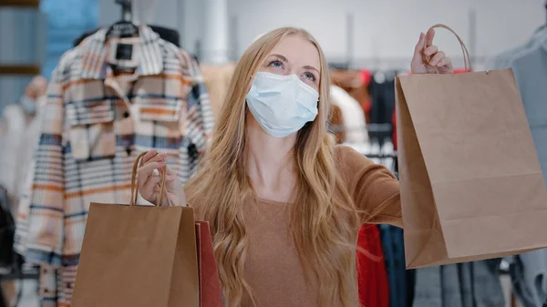 Rich happy blonde woman shopper wearing medical face protective mask girl customer client in clothing boutique with shopping bags dancing enjoying black friday discounts successful buying luxury cloth — 图库照片
