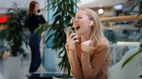 Caucasian funny blonde girl wears wireless headphones listens to music in mobile musical application online enjoys high-quality sound 30s woman sings song loudly dancing indoors with smartphone app — Stock Photo, Image