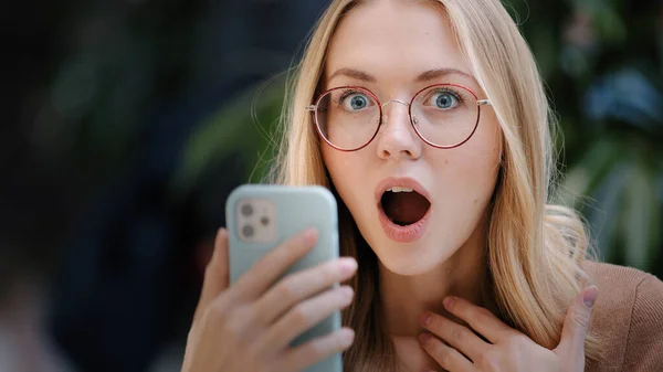 Portrait caucasian girl blonde successful lucky woman in glasses looks at screen of mobile phone wins online message with offer invitation happiness victory shock emotion good news with smartphone — Stok fotoğraf