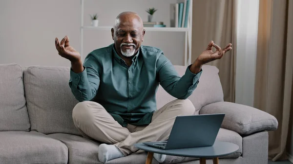 Calm mindful old 60s business man break sit at home on couch meditating breathing exercise feel no stress relief relaxing after work problem senior mature African male practicing yoga peaceful mind — Stock fotografie