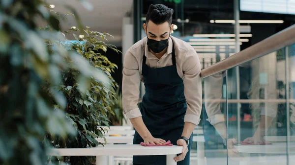 Arab man worker hispanic male waiter in medical mask catering worker in apron wipes table splashing with disinfectant does cleaning before opening restaurant prepares cafe for open visitors works — ストック写真