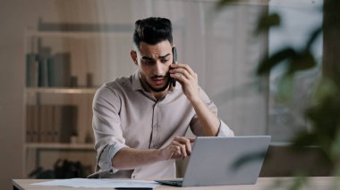 Bin yıllık Arap iş adamı web tasarımcısı cep telefonuyla konuşuyor çoklu görevlerde yer alan bilgisayarlar kullanarak online projeleri tartışıyor ofisteki kağıt dökümanlarını not ediyor.