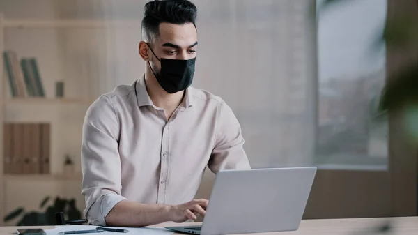 Hispanic arab male business man masked worker programmer typing on laptop search online information in cyberspace developing corporate project using software app chatting during coronavirus quarantine — стоковое фото