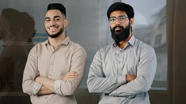 二つの幸せな男性同僚多人種多様なビジネスパートナー男性ヒスパニック男性アラビア語ひげを生やした男笑顔ポージングでオフィスでクロス腕を見ながらカメラに自信のある企業パートナーシップ — ストック写真
