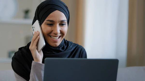 Felice donna araba sorridente ragazza musulmana in hijab donna freelance parlando dal telefono cellulare con l'amico che risponde chiamata godere di buone notizie ha negoziazione di affari utilizzare laptop vittoria online affare di successo — Foto Stock