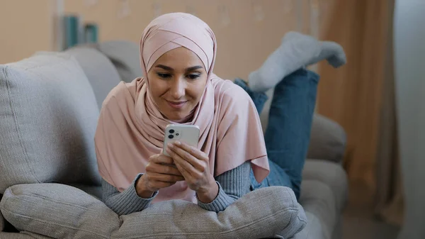 Giovane attraente araba musulmana donna islamica sdraiata sul divano di casa felice libanese signora indossare hijab rilassante digitando messaggio sul telefono cellulare con un amico fidanzato a guardare divertenti video shopping online — Foto Stock