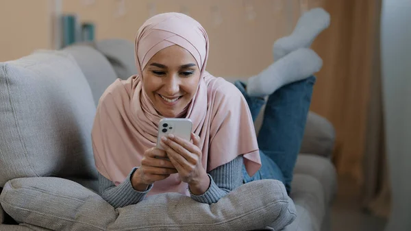 Young attractive arabian muslim girl islamic woman lying on home sofa happy lebanese lady wear hijab relaxing typing message on mobile phone with friend boyfriend watching funny video shopping online — Foto Stock