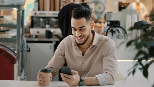 Indiase Spaanse Arabische man multiraciale zakenman zitten aan cafe tafel het drinken van hete koffie thee uit papieren beker op zoek in mobiele telefoon typen smartphone chatten browsen met behulp van online app gratis wi-fi — Stockfoto