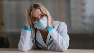 Yorgun portre, yorgun kız hemşire, doktor, kardiyolog, göz doktoru, jinekolog, stres basıncının masada oturan gözlüklü, koruyucu yüz maskesi taktığını düşünüyor.