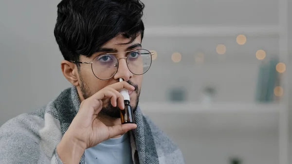 Close-up arabian indian bearded guy with glasses lonely sad sick ill man wrapped in blanket spraying rhinitis spray anti-allergic remedy drug in nose cold medicine seasonal allergy flue symptoms — 스톡 사진