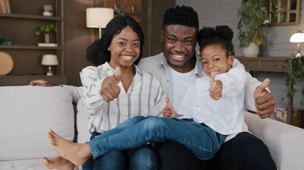 Happy African rodina mladí opatrovníci rodiče a malá dcera adoptované dítě sedí na pohovce doma vlastní byt nový dům při pohledu na foťák show palce nahoru schvalují pozitivní postoj — Stock fotografie