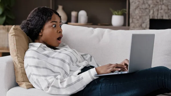 African girl freelancer american biracial woman lying on couch at home looking in laptop reading unexpected bad news gossip receiving notification opens mouth wide in surprise shock looking to camera