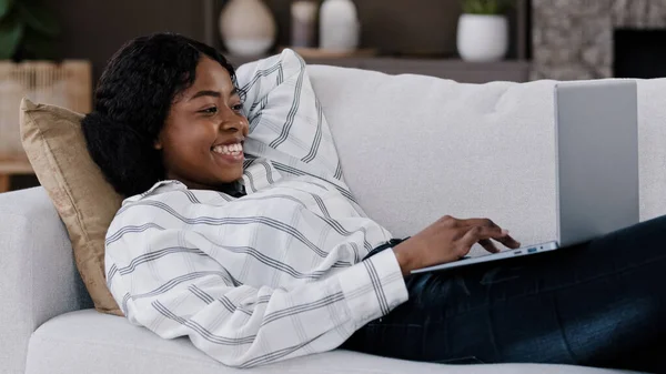 Sorridente donna africana studente ragazza freelance utente sdraiato sul divano utilizzando laptop digitando messaggio chat online con gli amici felice chat sul computer navigazione internet social media lavoro freelance — Foto Stock