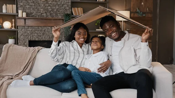 African couple sitting on couch under cardboard roof smiling looking at camera active happy daughter girl running sits on sofa. Relocation day to new home moving to own apartment homeowner portrait — Foto Stock