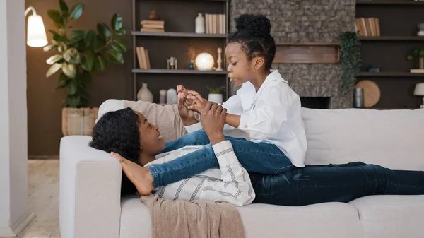 African American loving mother mommy lying on sofa relaxing at home living room with little daughter child talking having fun casual game playing holding hands baby rocking back forward sitting on mum — Foto Stock