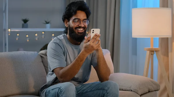 Ευτυχισμένος αραβικός ινδός άνδρας χιλιετίας κρατώντας σύγχρονο μήνυμα SMS smartphone στο σπίτι κάθεται στον καναπέ το βράδυ γενειοφόρος τύπος με γυαλιά χρησιμοποιώντας το κινητό app βλέποντας αστείο βίντεο σε απευθείας σύνδεση γέλιο — Φωτογραφία Αρχείου