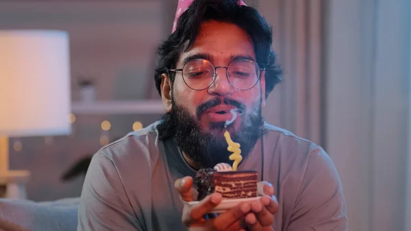 Porträtt arabiskt glad indian man ansikte skäggig man bär festlig födelsedag rosa hatt och glasögon håller cupcake gör önskan glad gratulera hemma blåser brinnande ljus på tårta fira fest — Stockfoto