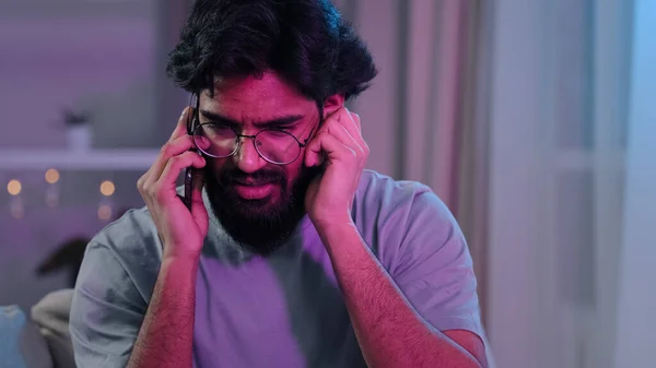 Arabic Indian bearded man with glasses talking on phone at house party sitting on sofa neon light hard to hear conversation loud music noisy birthday celebration answering phone call trying listening — Stock Photo, Image