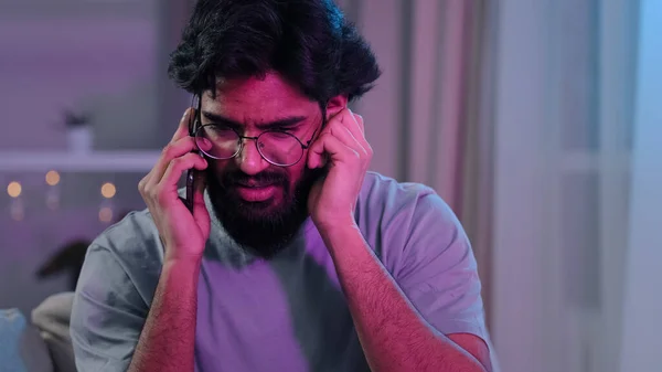 Árabe indio barbudo hombre con gafas hablando por teléfono en casa partido sentado en sofá neón luz difícil de escuchar conversación música ruidosa cumpleaños celebración contestar llamada telefónica tratando de escuchar —  Fotos de Stock