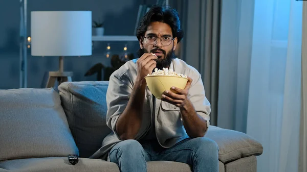 Serious interested Arabic Hispanic Indian bearded man guy wearing glasses 30s male with popcorn watching TV at home sofa late evening night relaxing enjoying movie online sport game attentively watch — Stockfoto